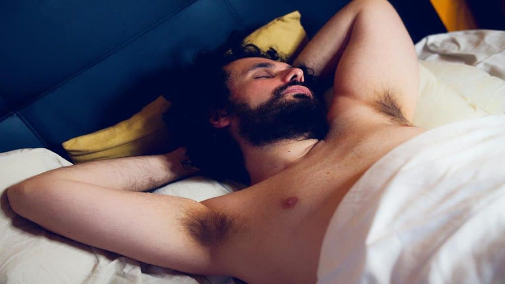 A bearded man sleeping shirtless in bed.