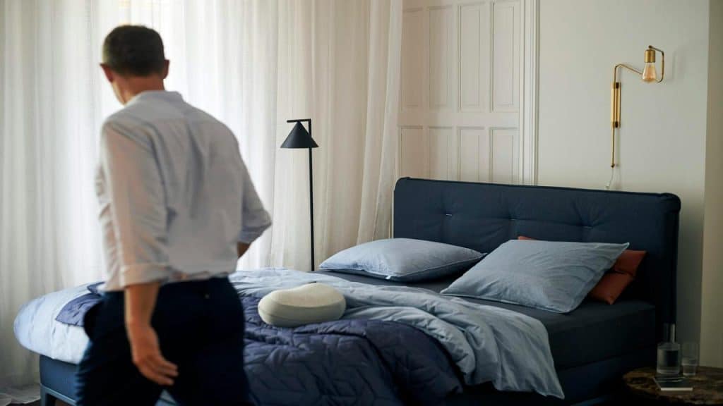 A man walking toward a neatly made bed in a softly lit bedroom.