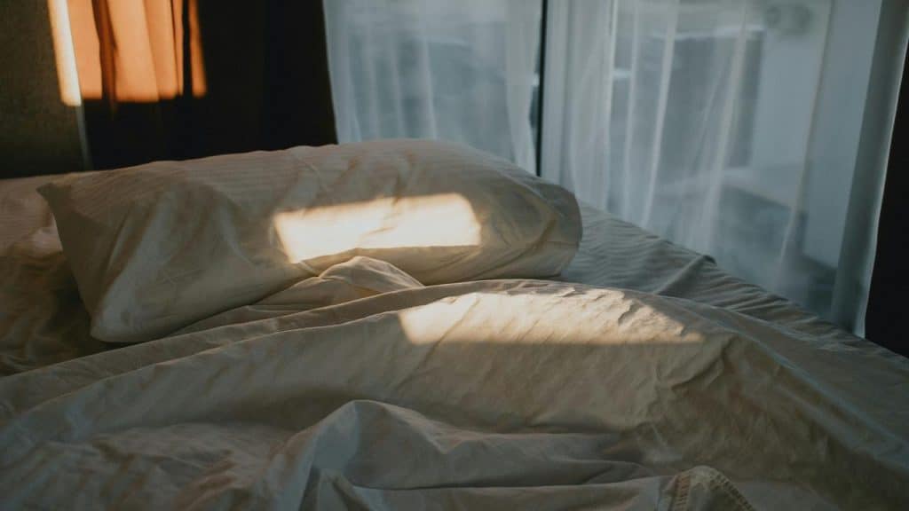A sunlit bed with wrinkled white sheets and a single pillow near a window with sheer curtains.