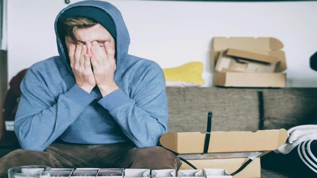 A man in a hoodie sitting on a couch, covering his face with his hands in frustration or stress.