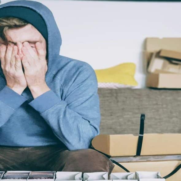 A man in a hoodie sitting on a couch, covering his face with his hands in frustration or stress.