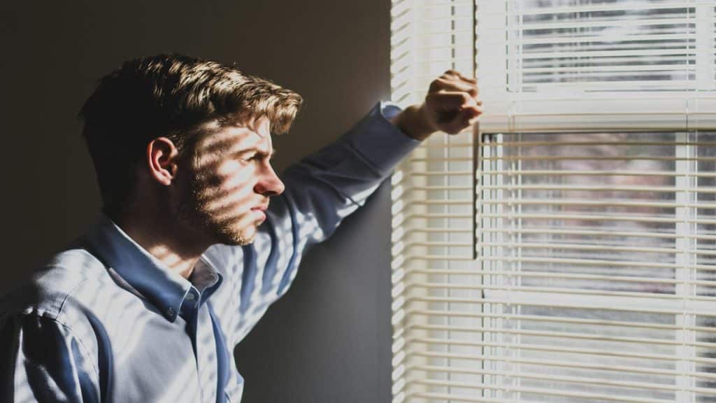 A man standing by a window with blinds, resting his arm on the frame, looking outside pensively.