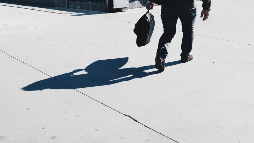 A man walking away while carrying a bag, his long shadow cast on the ground in the sunlight.