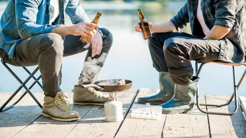 A relaxed moment as two men sit on a dock, grilling food and drinking beer by the water.