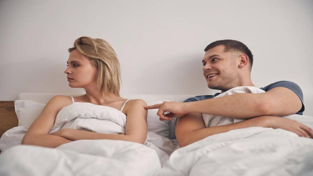 A couple in bed where the woman looks away as the man smiles and playfully pokes her.