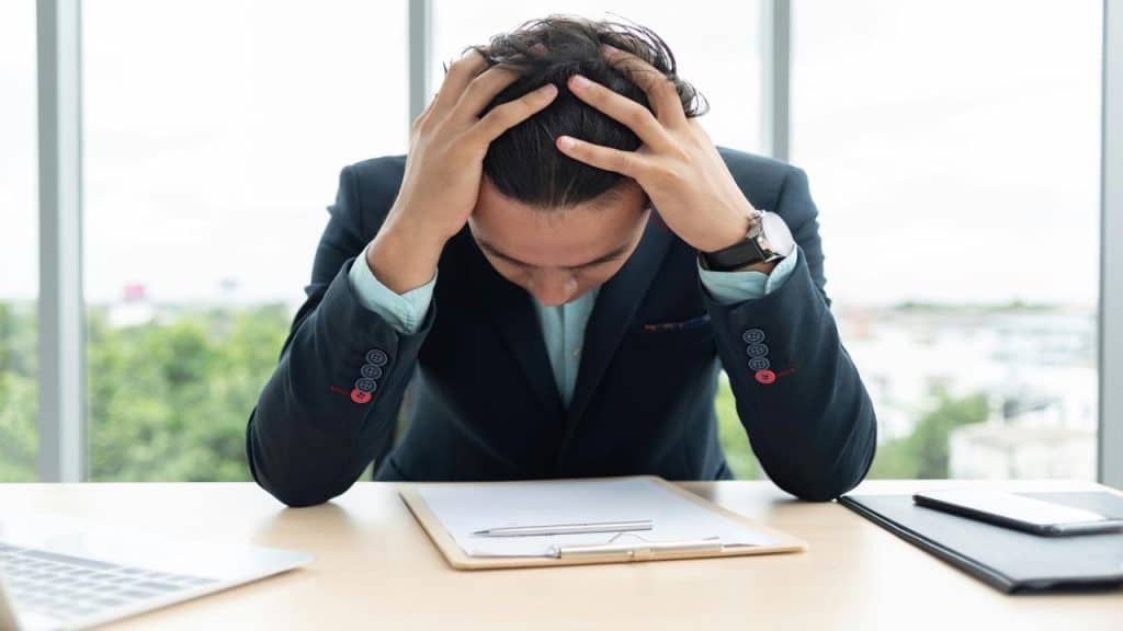 A man in a suit sits at a desk with his head down and hands gripping his hair, looking stressed.