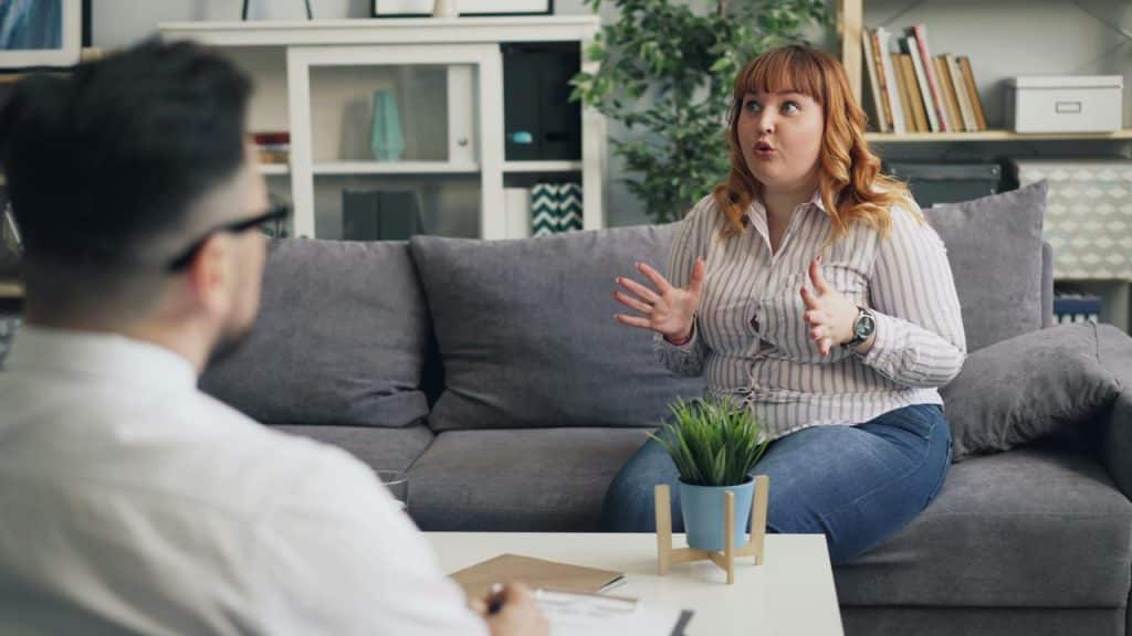 A woman sits on a couch speaking animatedly to a man in front of her.