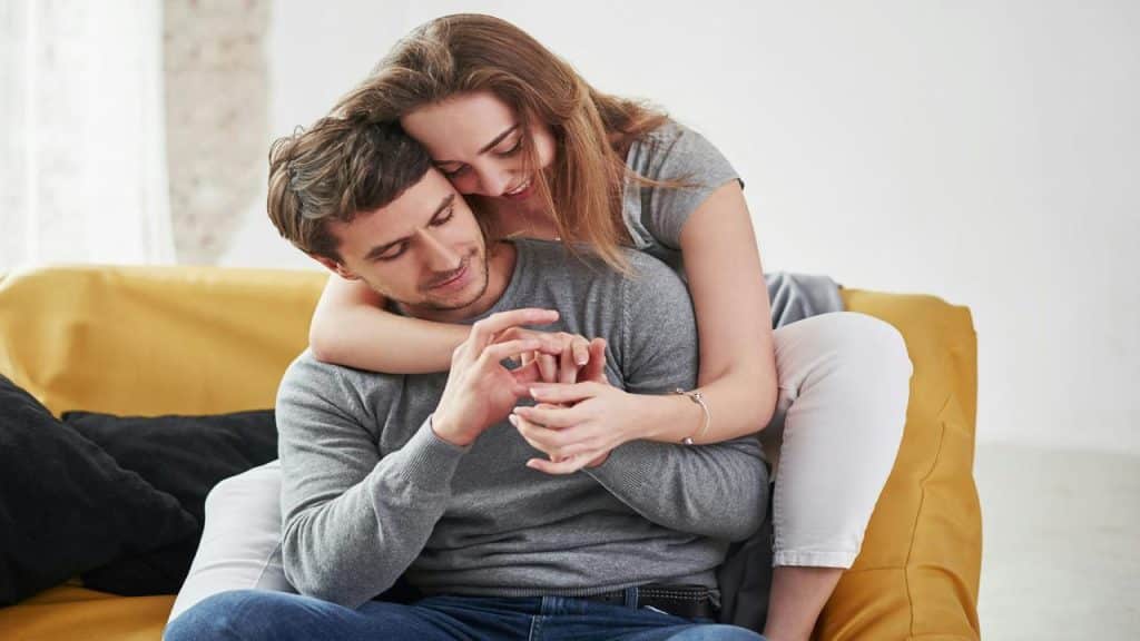A woman embraces a man from behind on a couch, both smiling as she places a ring on his finger.