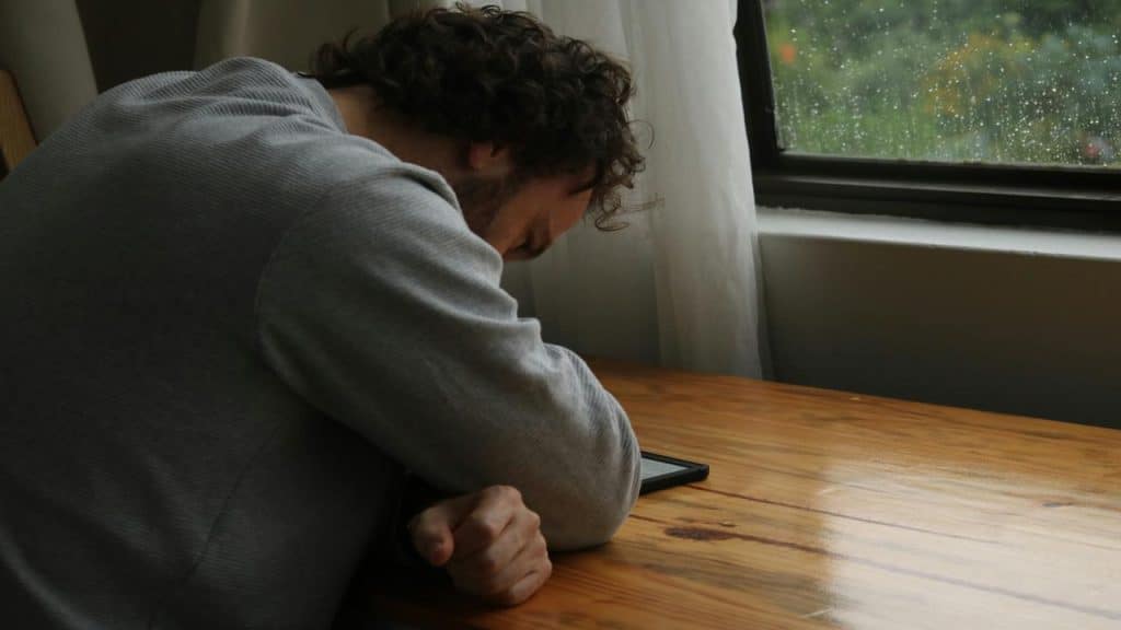 A man sits at a wooden table with his head resting on his arm, looking down at a phone, next to a rainy window.