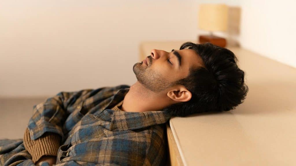 A man leans back against a counter with his eyes closed, wearing a plaid jacket, appearing calm or deep in thought.