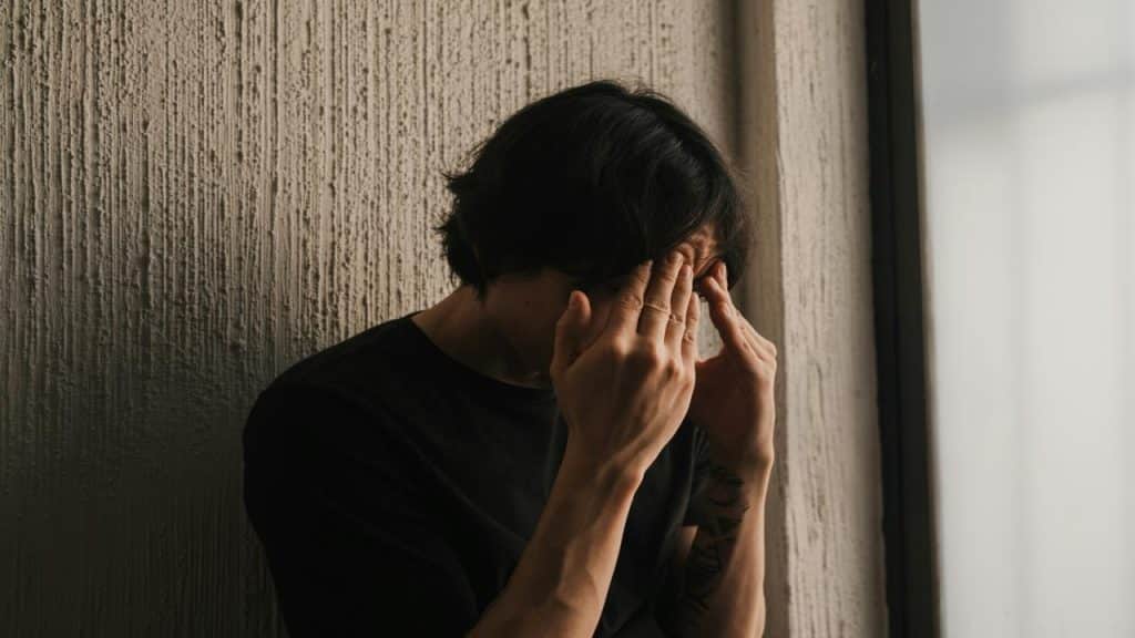 A man in black leans against a wall covering his face.