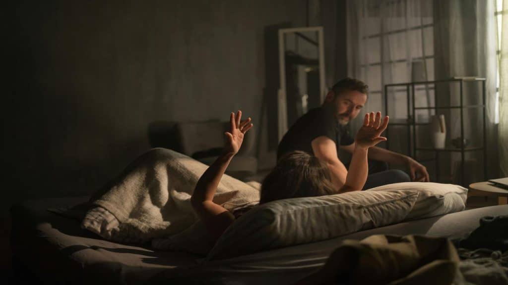 A woman lies in bed with her arms raised while a man sits nearby looking at her.