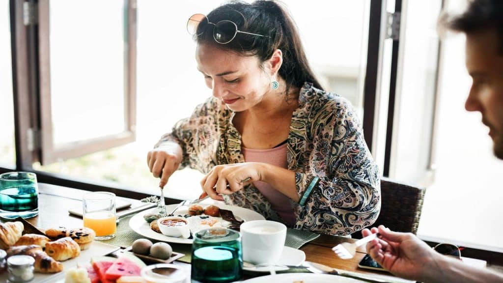 A woman is enjoying a meal at a table, cutting her food while smiling, with a variety of breakfast dishes and drinks.
