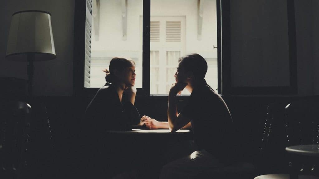 A couple sits at a table, holding hands and gazing at each other in a softly lit room by a window.