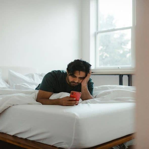 A man lying on a bed with white sheets, looking at a phone in his hand.