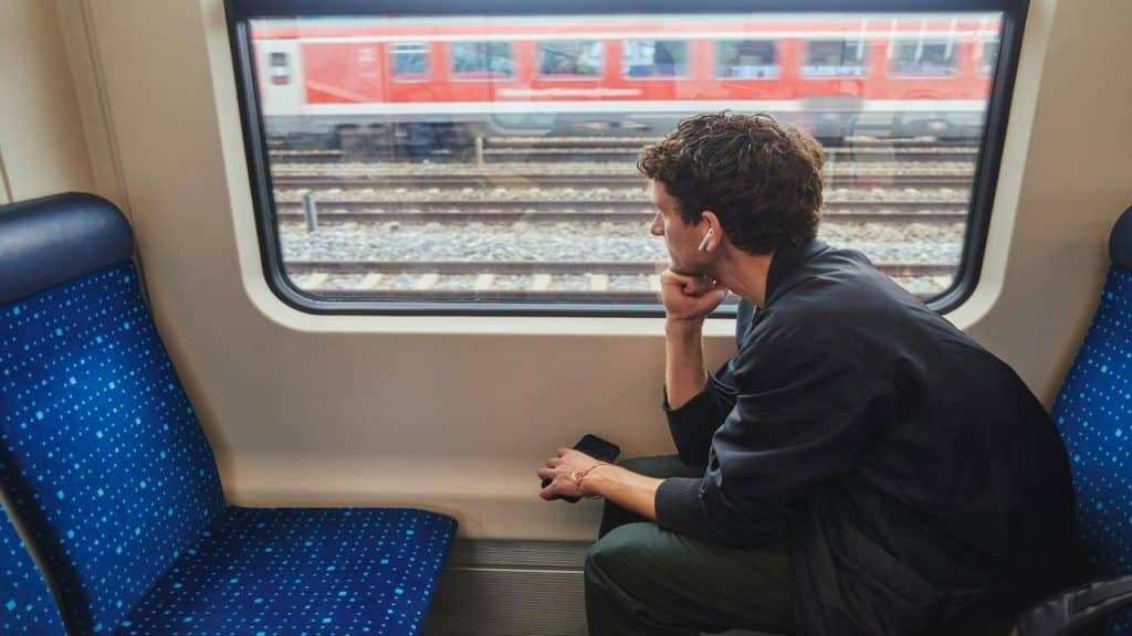 A man on a train, wearing earphones and holding a phone, gazing out the window at the tracks and a passing train.