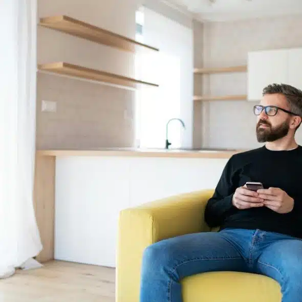 A man sitting on a yellow couch holding a phone, looking out the window.