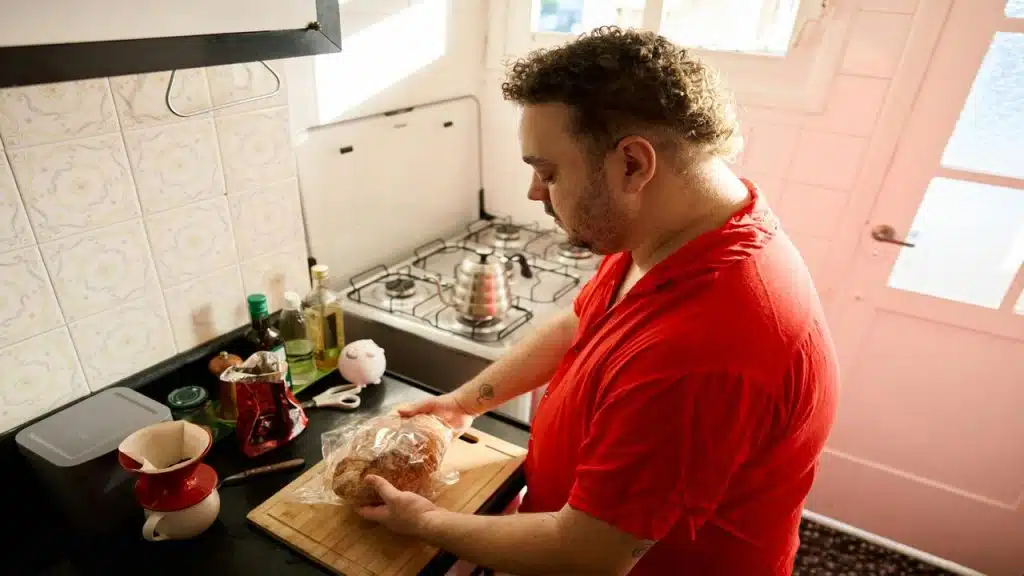 A person in a red shirt holding a bag of bread in a small kitchen.