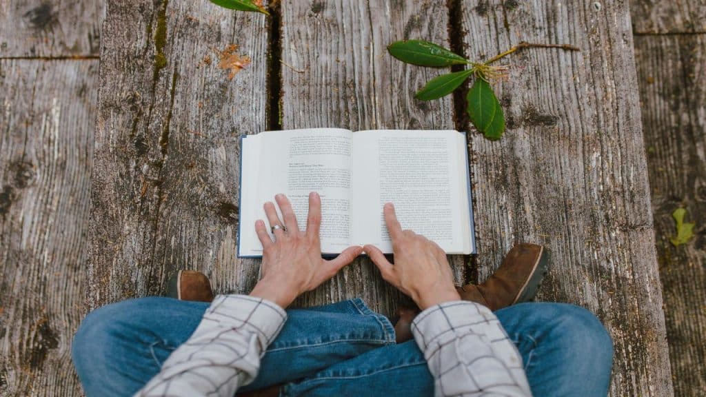 A person sitting cross-legged on wooden planks reading an open book.