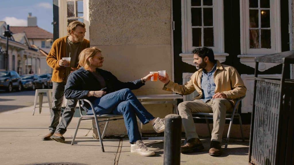 An outdoor scene of three men with drinks, two clinking cups.
