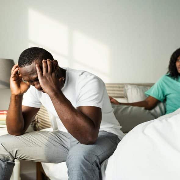 A frustrated man sitting on the edge of a bed with a concerned woman talking behind him.