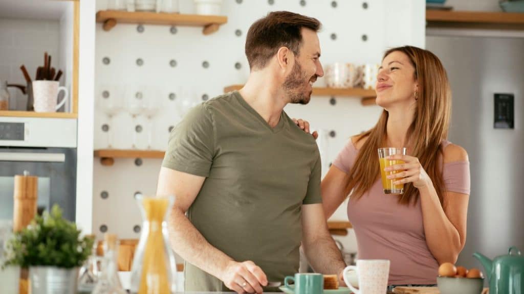A man and a woman in a kitchen smile at each other, with the woman holding a glass of juice.