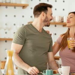A man and a woman in a kitchen smile at each other, with the woman holding a glass of juice.