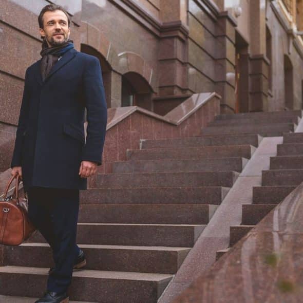 A well-dressed man carrying a brown duffel bag walks down a set of stone steps.