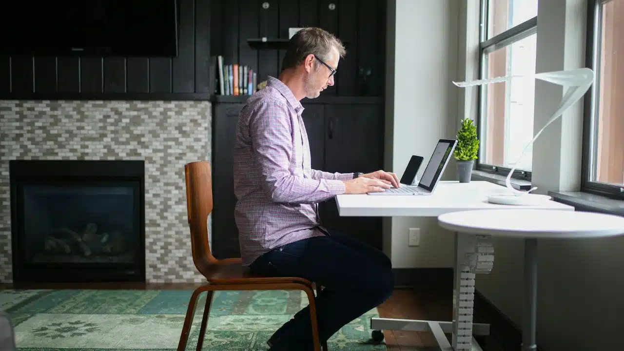 A man wearing glasses and a plaid shirt works on a laptop at a white desk.