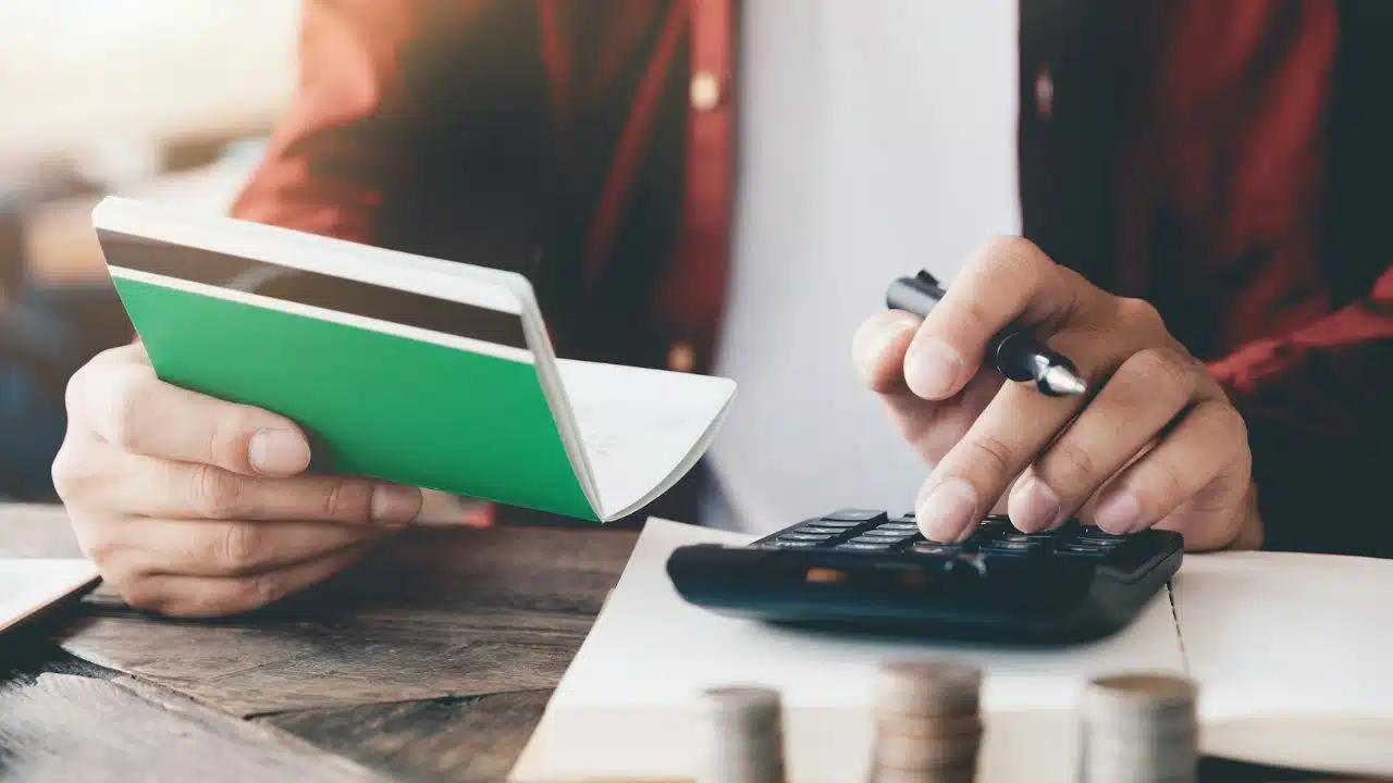 A person holds a green passbook while using a calculator, with coins stacked nearby.