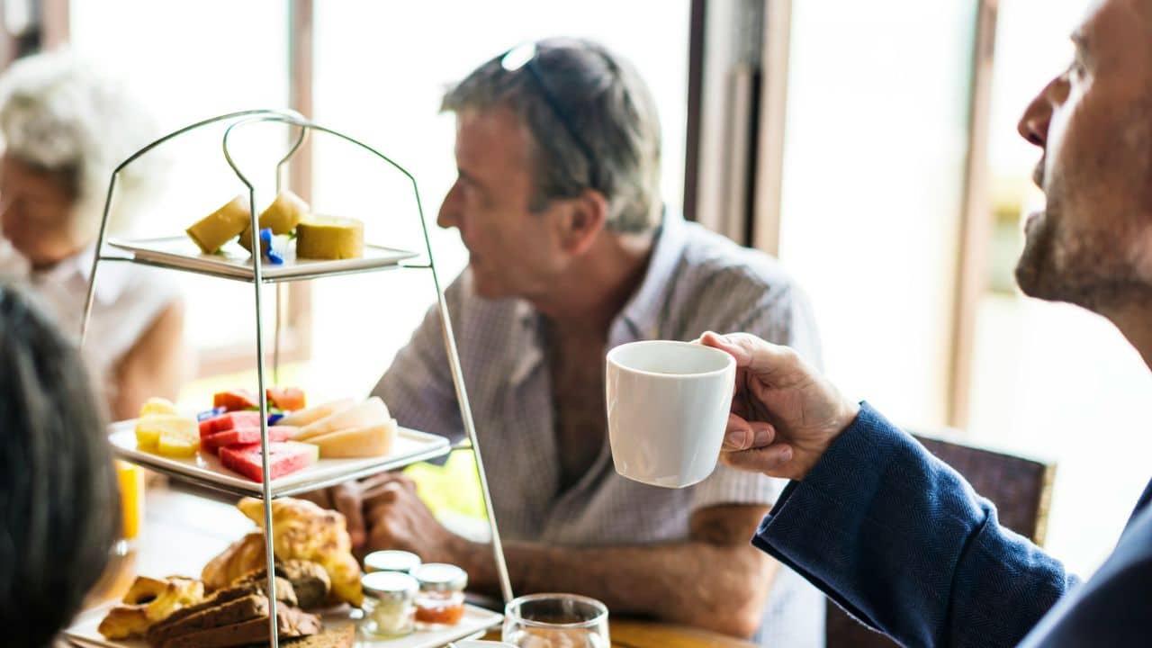 A person holds a white cup while sitting at a table with a tiered tray of snacks.