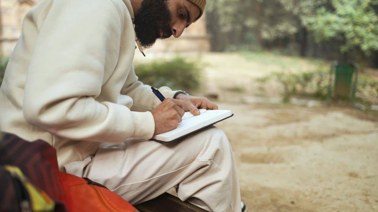 A bearded man wearing a beanie sits outdoors, writing in a notebook.
