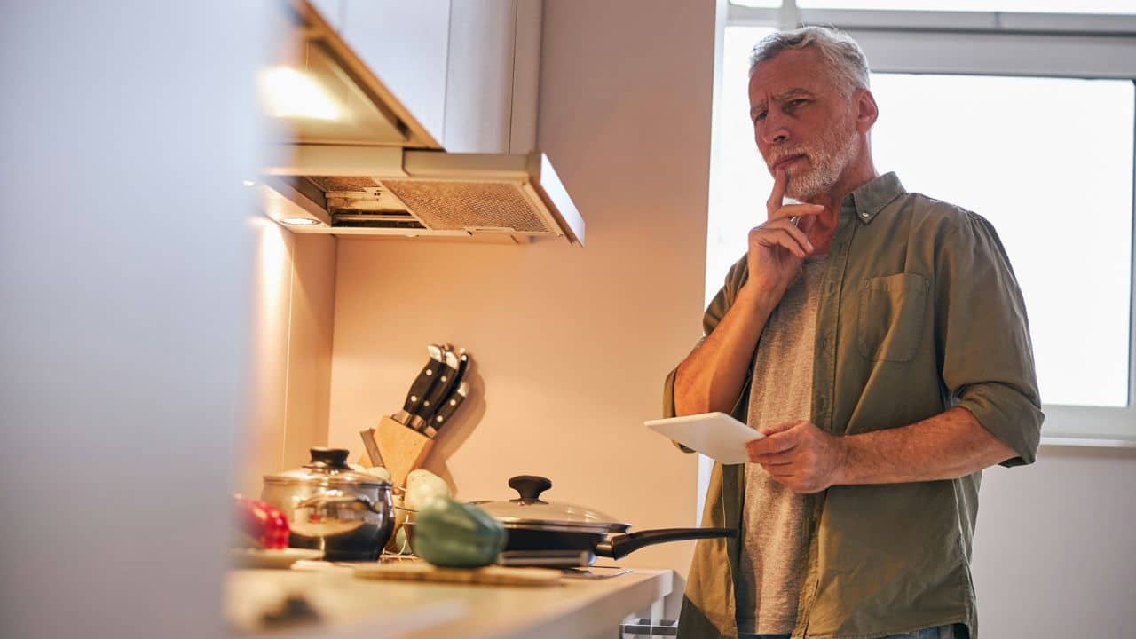 A bearded man in a kitchen holds a tablet and looks thoughtful, with pots on the counter.