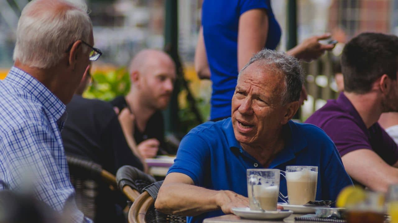 A man in a blue polo shirt talks at an outdoor cafe table with two coffee drinks.