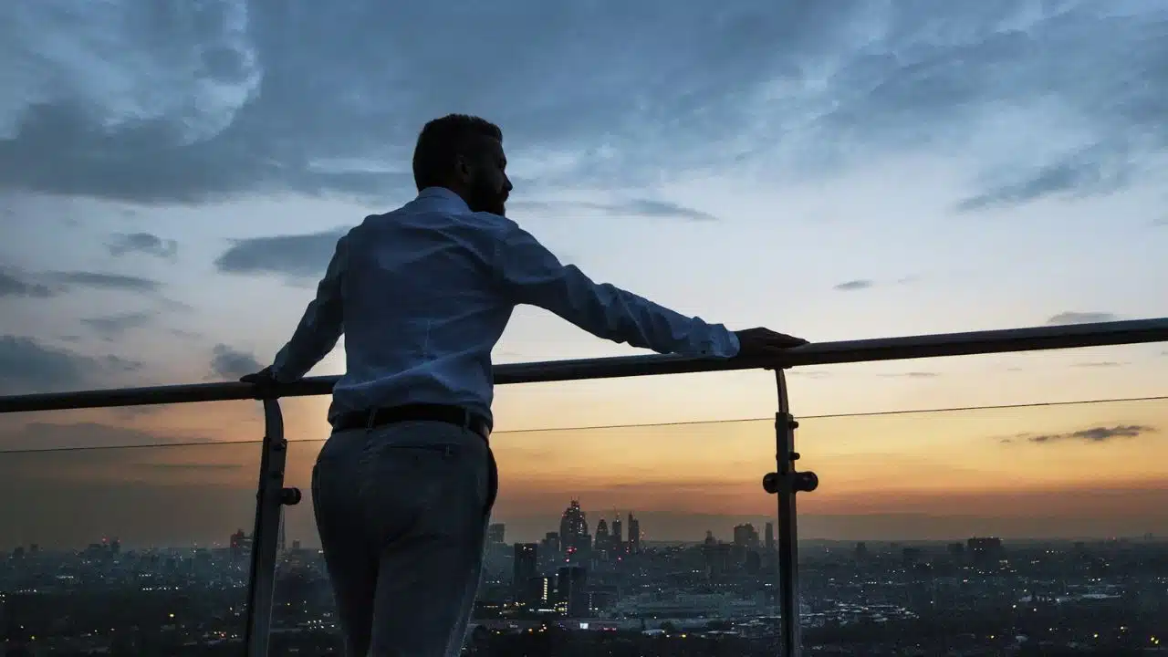 A man in a white shirt and light pants leans on a railing, looking at a city skyline at dusk.