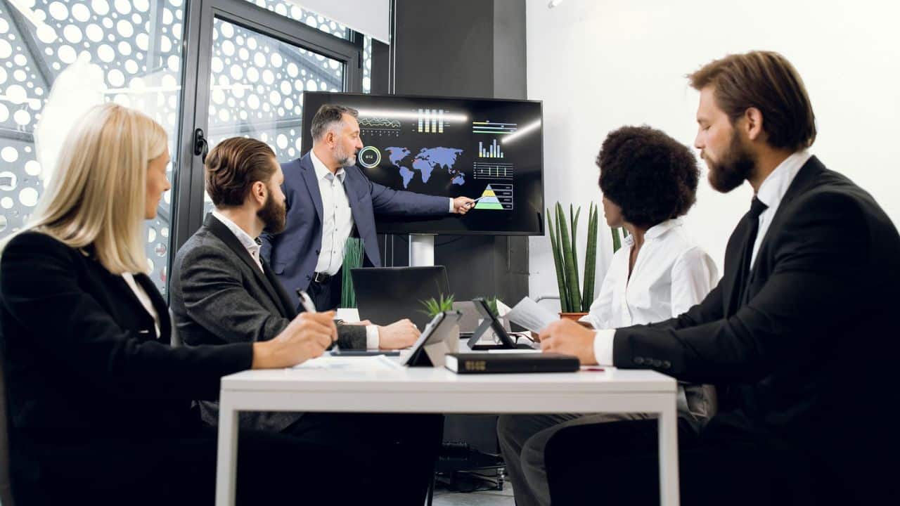 A diverse group of five business professionals is engaged in a meeting, with one man pointing at a large screen displaying charts and a world map.