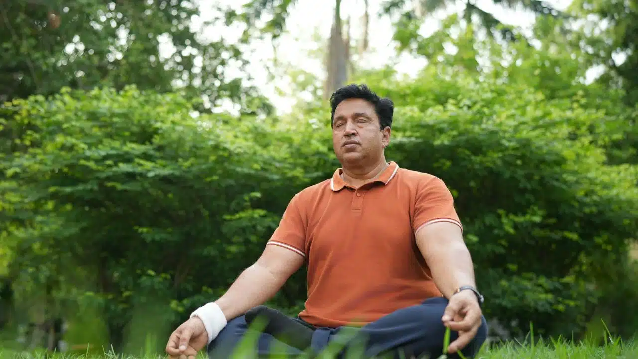 A man in an orange polo shirt sits cross-legged in a grassy area with his eyes closed.
