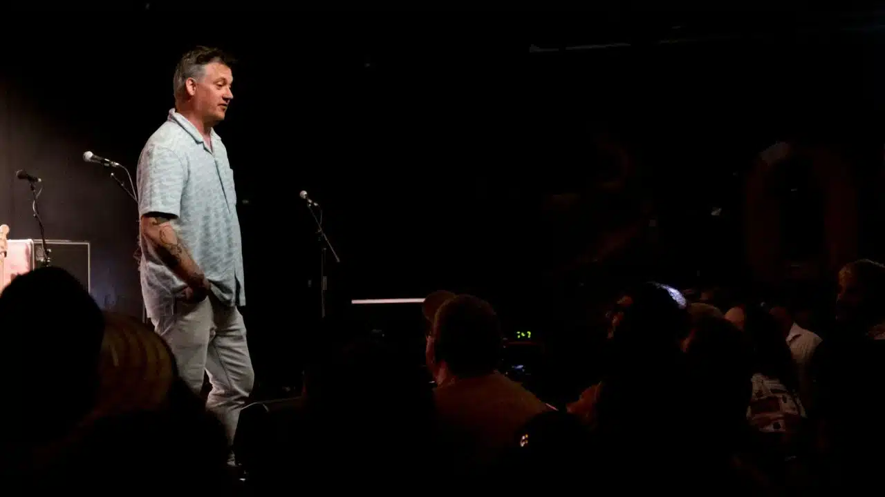A man in a light shirt stands on a stage with microphones, facing a dark audience.