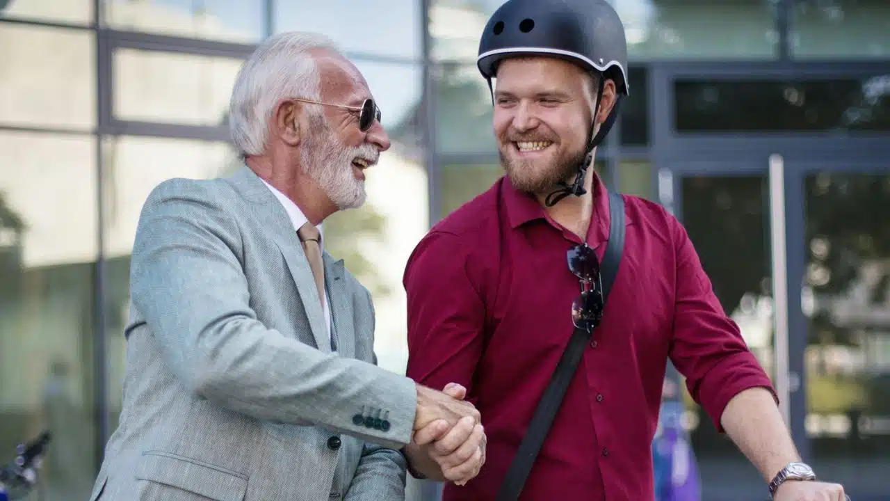 A senior man in a suit shakes hands with a younger man wearing a helmet and a red shirt.