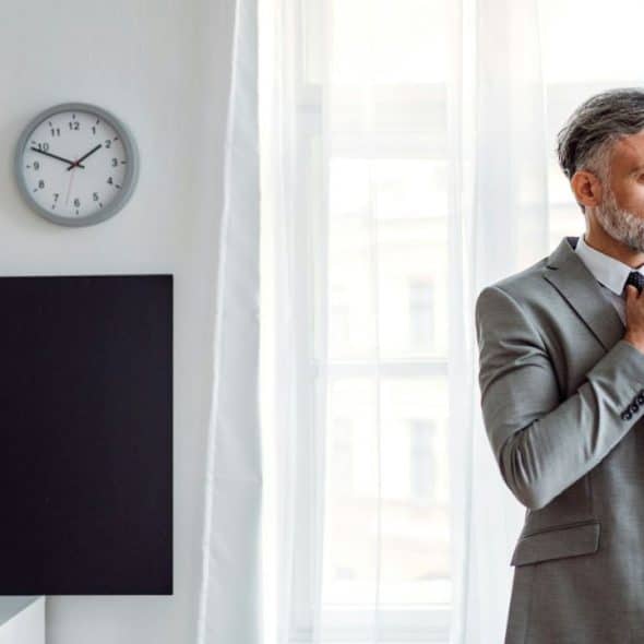 A distinguished man in a grey suit adjusts his tie, looking out a window.
