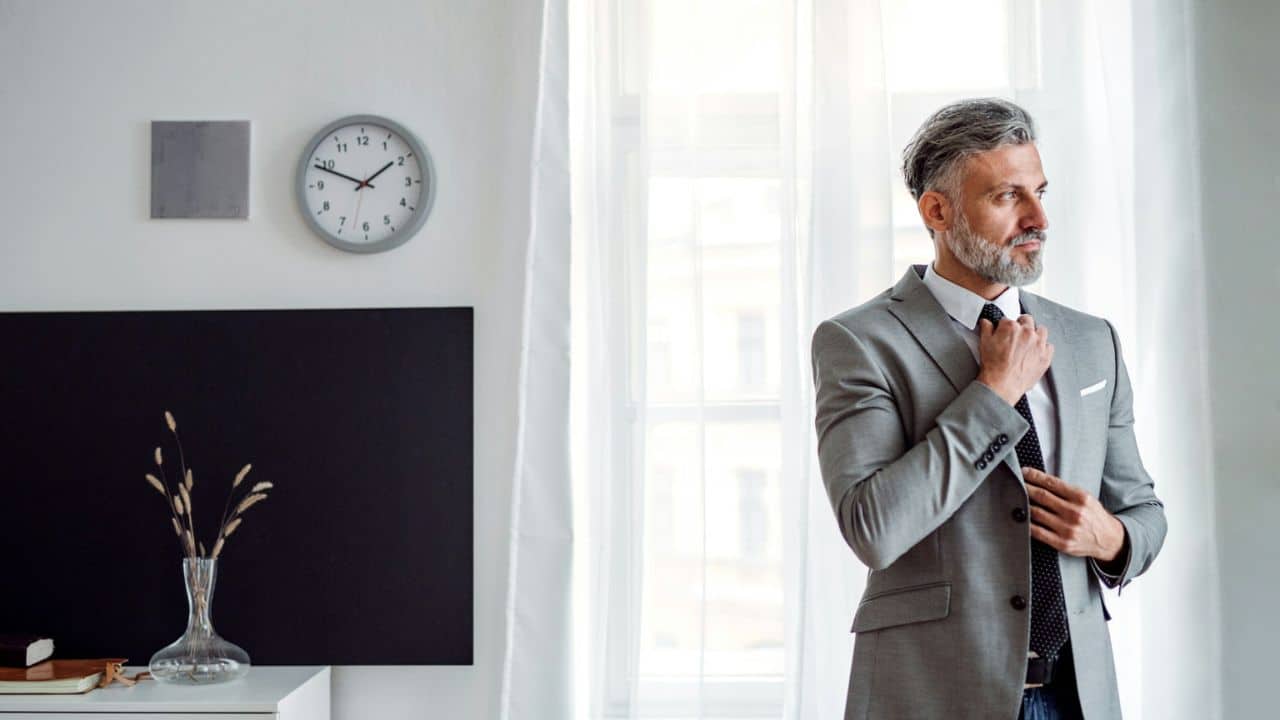 A distinguished man in a grey suit adjusts his tie, looking out a window.