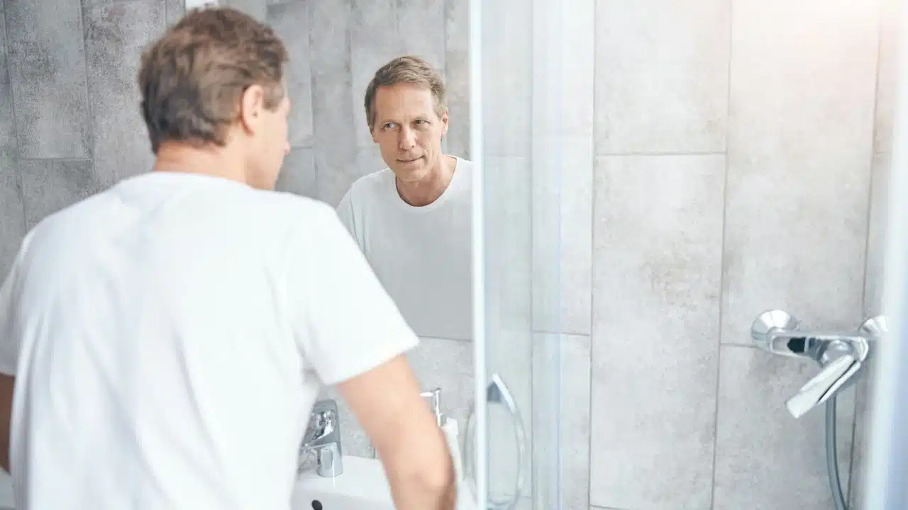 A man in a white t-shirt looks at his reflection in a bathroom mirror.
