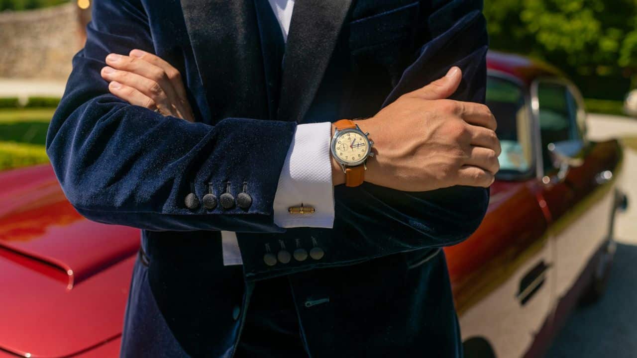 A man in a blue velvet suit with crossed arms wears a watch and cufflinks, standing by a red car.