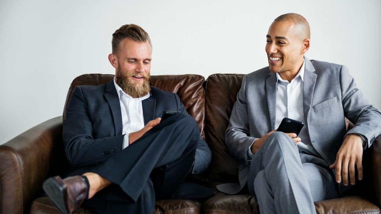 Two men in suits sit on a brown leather couch, smiling and holding phones.