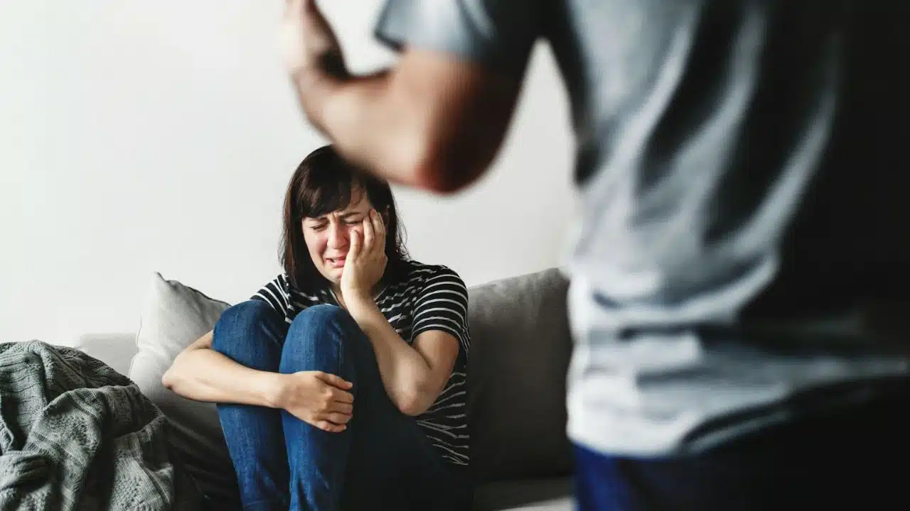 A distressed woman sits on a couch, crying, with a man's back to the viewer in the foreground.