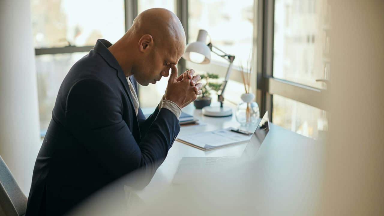 A bald man in a suit sits at a desk with his head in his hands, appearing stressed.