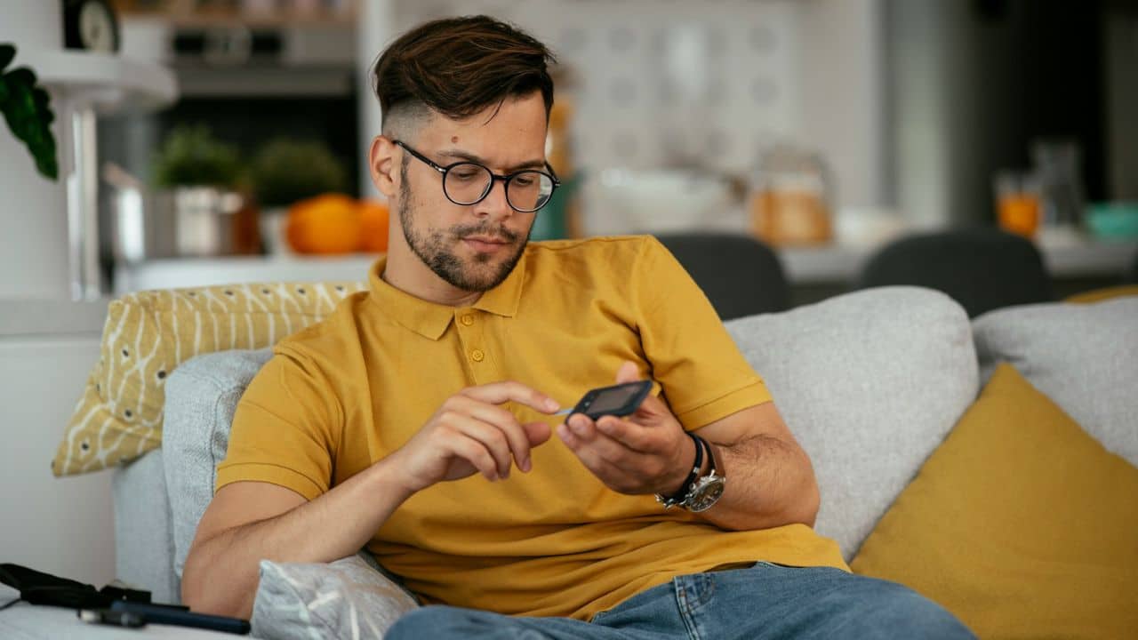 A man with glasses and a beard sits on a sofa, looking at his phone.