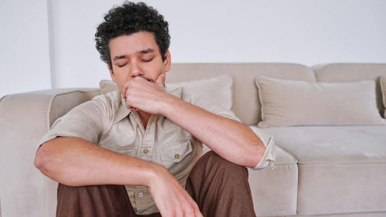 A man with curly hair and a mustache sits on the floor, eyes closed, with his hand on his chin.