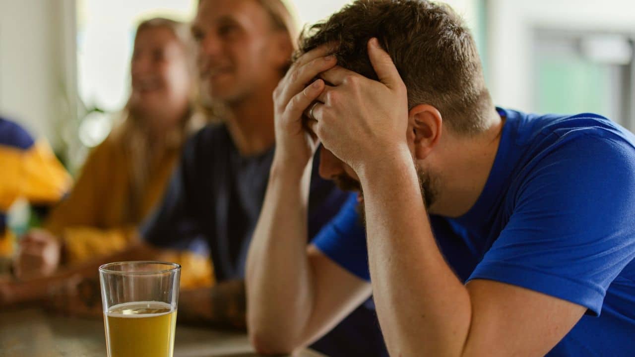 A man in a blue shirt holds his head in his hands, with a beer glass in the foreground.