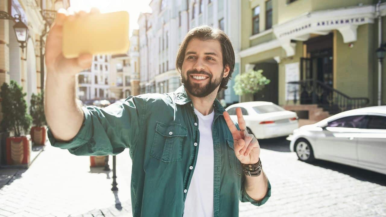 A smiling man with a beard takes a selfie on a sunny city street.
